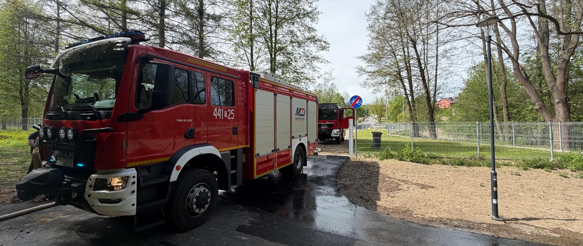 Dwa czerwone wozy strażackie stoją na mokrej drodze przy parku; wokół rosną drzewa, widać ogrodzenie i latarnię, a na jezdni są ślady wody po akcji gaśniczej.