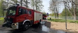Dwa czerwone wozy strażackie stoją na mokrej drodze przy parku; wokół rosną drzewa, widać ogrodzenie i latarnię, a na jezdni są ślady wody po akcji gaśniczej.