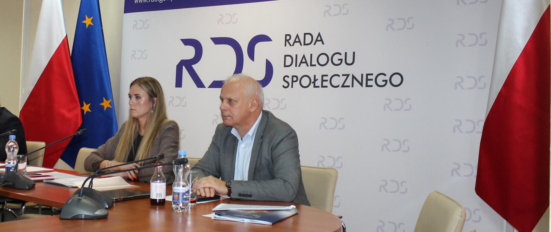 Zdjęcie przedstawia stół prezydialny za którym siedzą: Kamila Król - Podsekretarz Stanu w Ministerstwie Rozwoju i Technologii, Współprzewodnicząca Zespołu ze strony rządowej, Andrzej Karol - Współprzewodniczący Zespołu ze strony związków zawodowych. W tle siedzących znajduje się baner z napisem Centrum Partnerstwa Społecznego Dialog i. Andrzeja Bączkowskiego. Po lewej i prawej stronie zdjęcia stoją po dwie flagi: Polska i UE. Nad banerem widnieje Godło Polski.