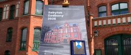 Tablica informacyjna konkursu " Zabytek Zadbany 2026" i oprawiony dyplom na sztaludze przed ceglanym budynkiem Państwowej Szkoły Muzycznej I i II st. im. Oskara Kolberga w Szczecinku. 