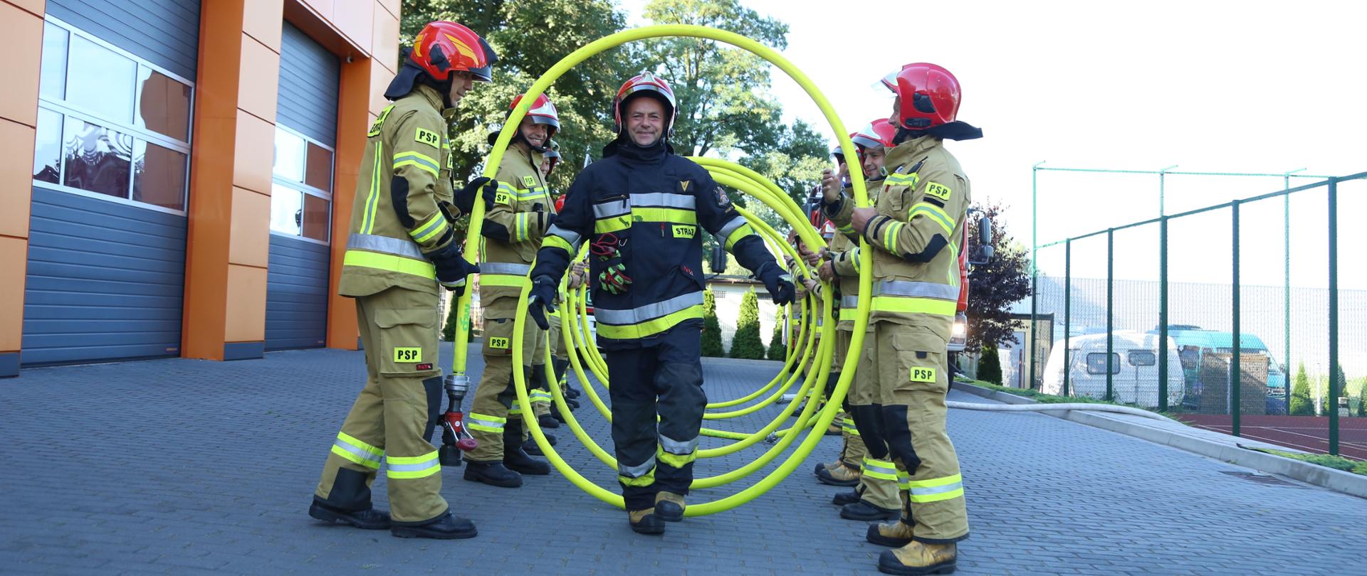 Strażacy stoją w dwóch szeregach, trzymają wąż, strażak przechodzi pośrodku