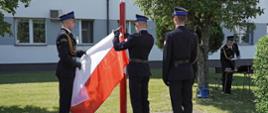 . trzech strażaków ubranych w mundury galowe, jeden trzyma flagę państwową, drugi stoi w pozycji na baczność a trzeci mocuje flagę do masztu, w tle budynek komendy straży pożarnej, rosną dwa drzewa a pod jednym z nich stoi strażak ubrany w mundur galowy, pogoda słoneczna