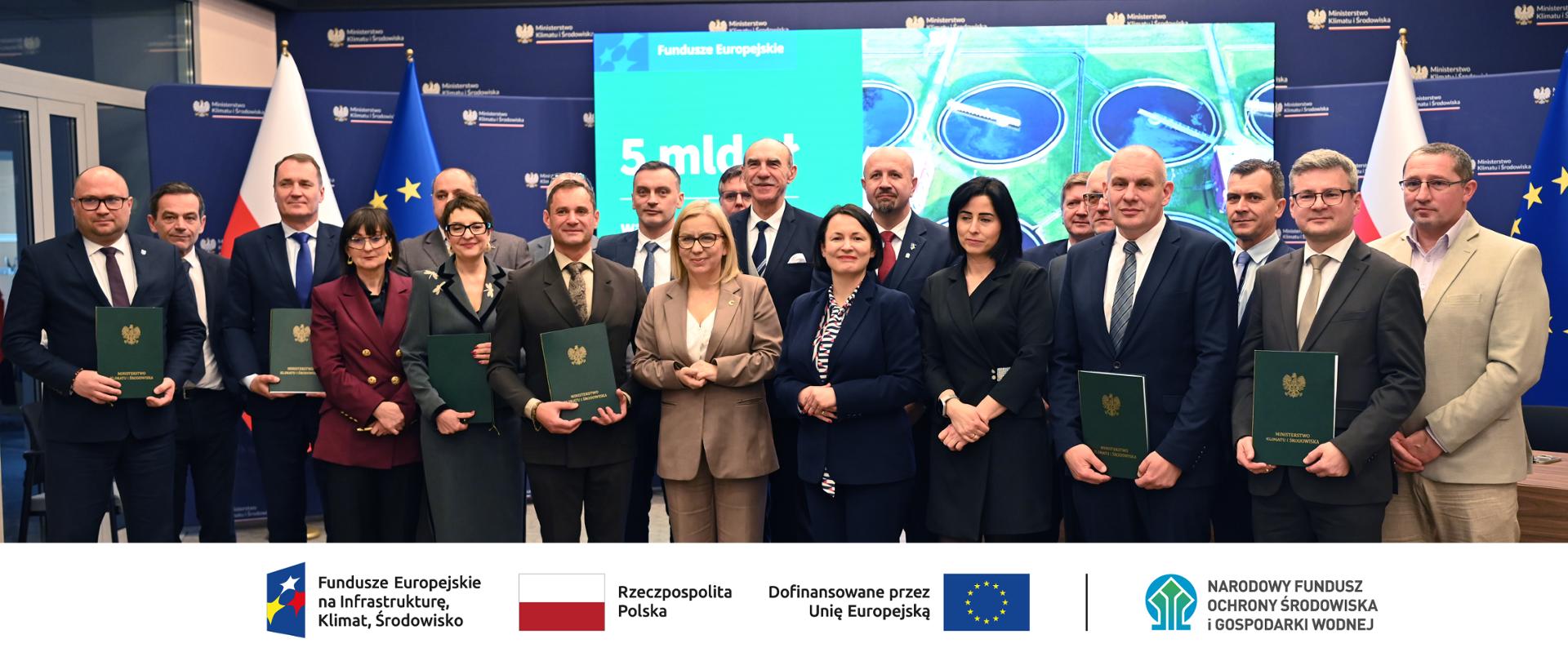 Zdjęcie grupowe. W stopce grafiki ciąg logotypów: Fundusze Europejskie na Infrastrukturę, Klimat, Środowisko, flaga Rzeczypospolita Polska, flaga UE Dofinansowane przez Unię Europejską, Narodowy Fundusz Ochrony Środowiska i Gospodarki Wodnej.