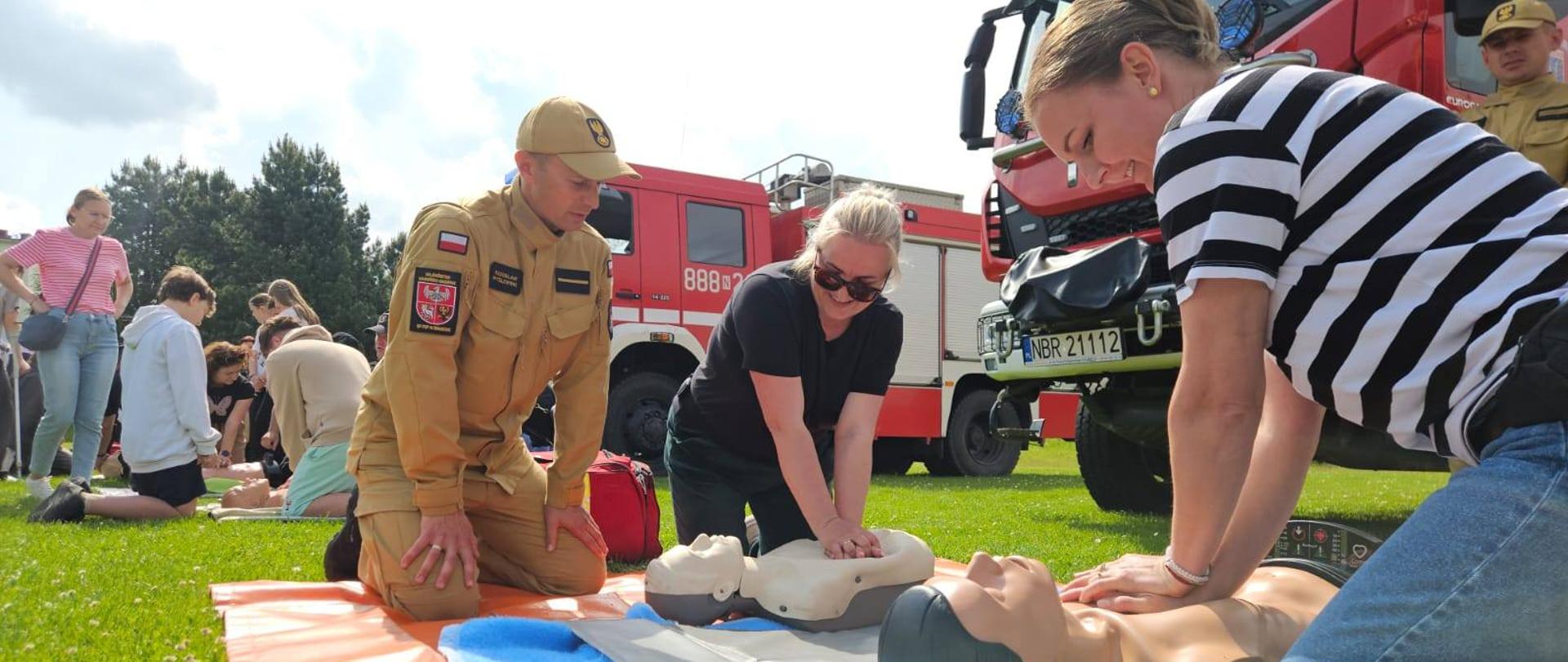 na zdjęciu z prawej strony dwie kobiety które uciskają manekina, przy nich strażak. Trwa pokaz pierwszej pomocy. W tle samochody strażackie oraz inne osoby. Słonecznie.