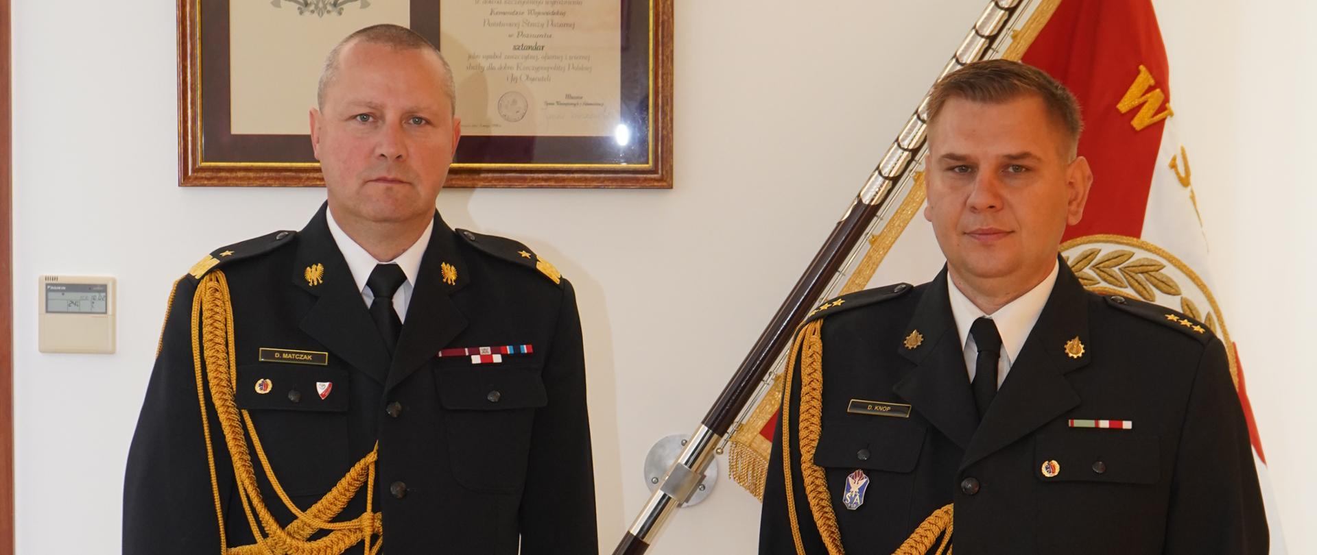 dwóch mężczyzn w mundurach strażackich ze sznurem stoi na baczność, jeden trzyma w ręku teczkę, w tle sztandar