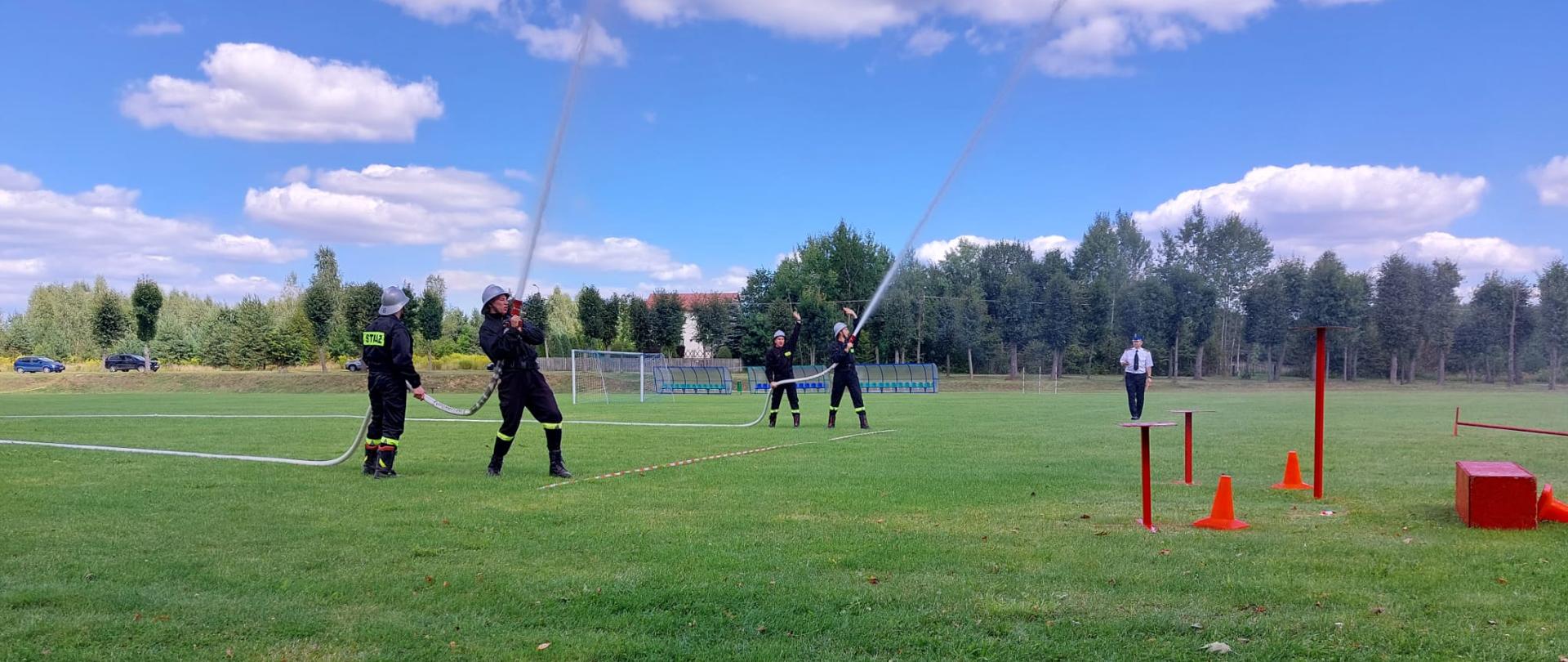 Na zielonym boisku dwie roty strażaków podają do góry prądy wody. 