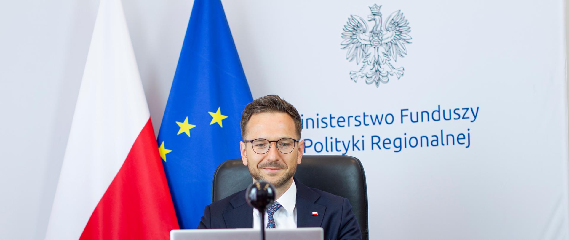 Wiceminister Waldemar Buda siedzi przy biurku, przed laptopem. Za plecami tło z logo MFiPR oraz flagi Polski i UE.