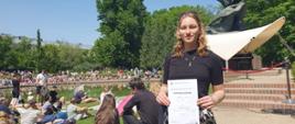 Na tle pomnika stoi uśmichnięta laureatka konkursu. W dłoniach trzyma dyplom. Jest piękny letni dzień. W otoczeniu drzew parku w Łazienkach widoczne grupki zgromadzonych słuchaczy koncertów chopinowskich.