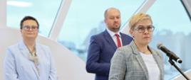 Pod półokrągłym oknem stoi wiceminister Lubnauer i dyrektor IBE w granatowym garniturze, przed nimi stoi wiceminister Mrówczyńska która mówi do mikrofonu na stojaku i trzyma w ręku czerwoną teczkę.