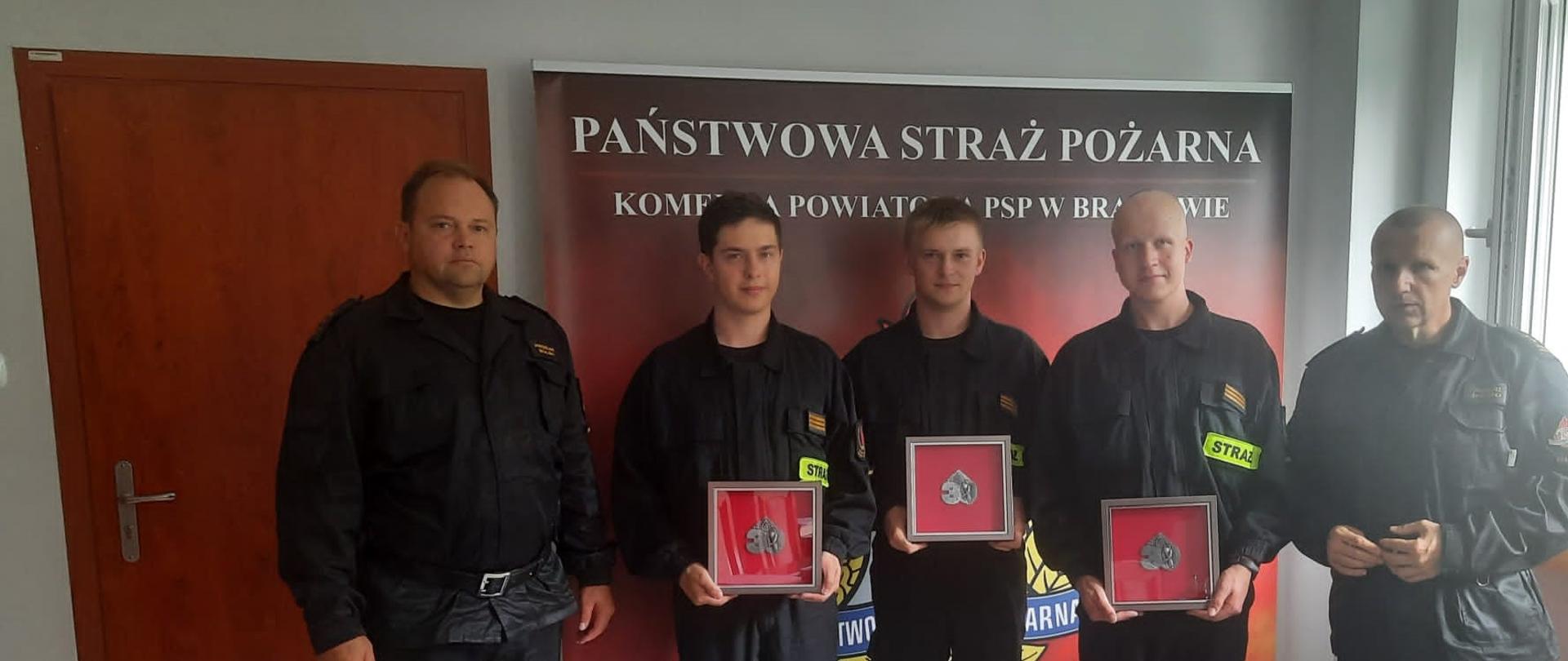 Z prawej strony w gabinecie stoi Komendant Powiatowy PSP, z lewej Zastępca komendanta PSP a po środku na tle baneru z logiem PSP trzech studentów w mundurach PSP z upominkami.