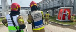 Na pierwszym planie dwóch strażaków Kierujących Działaniem Ratowniczym, z prawej strony komora dekontaminacyjna i obsługujący ją strażacy. W oddali reszta zakładu mleczarskiego Hochland.