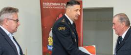 Komendant Powiatowy umundurowany w mundur galowy wręcza w asyście Posła na Sejm RP Decyzję o przyznaniu świadczenia ratowniczego druhowi OSP