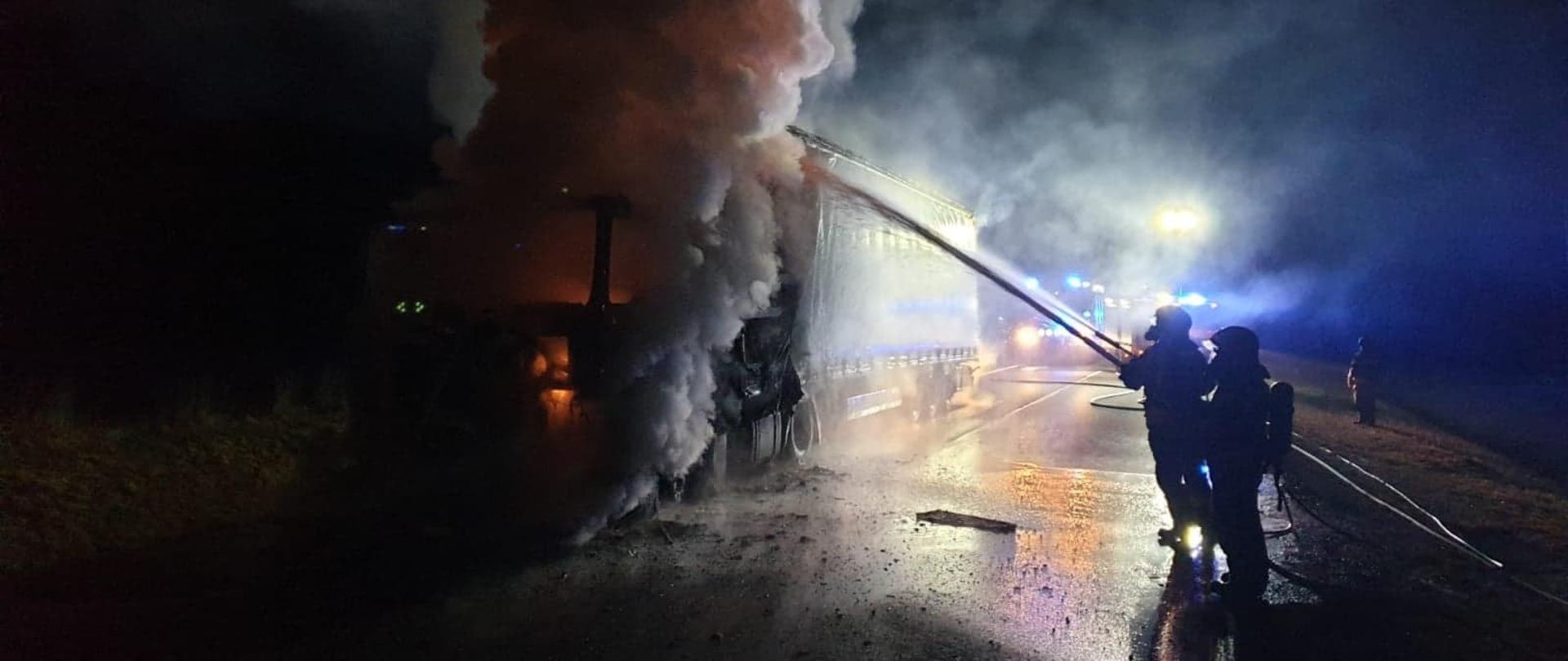 Strażacy podają prąd piany na palący się ciągnik siodłowy z naczepą stojący na jednym pasie drogi.