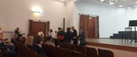 Na zdjęciu uchwycono moment wręczenia nagród podczas IX Ogólnopolskiego Konkursu Gitarowego Szkół Muzycznych I Stopnia „Gitarowe NasTroje”, który odbył się 11 czerwca 2026 roku w Warszawie. W centralnej części kadru znajduje się oficjalny moment odbioru wyróżnienia — uczeń naszej szkoły odbiera nagrodę za zajęcie II miejsca w konkursie. Wokół stoją organizatorzy i uczestnicy wydarzenia, a na widowni siedzą uczniowie, opiekunowie i goście obserwujący ceremonię. Po prawej stronie widać scenę przygotowaną do występów.