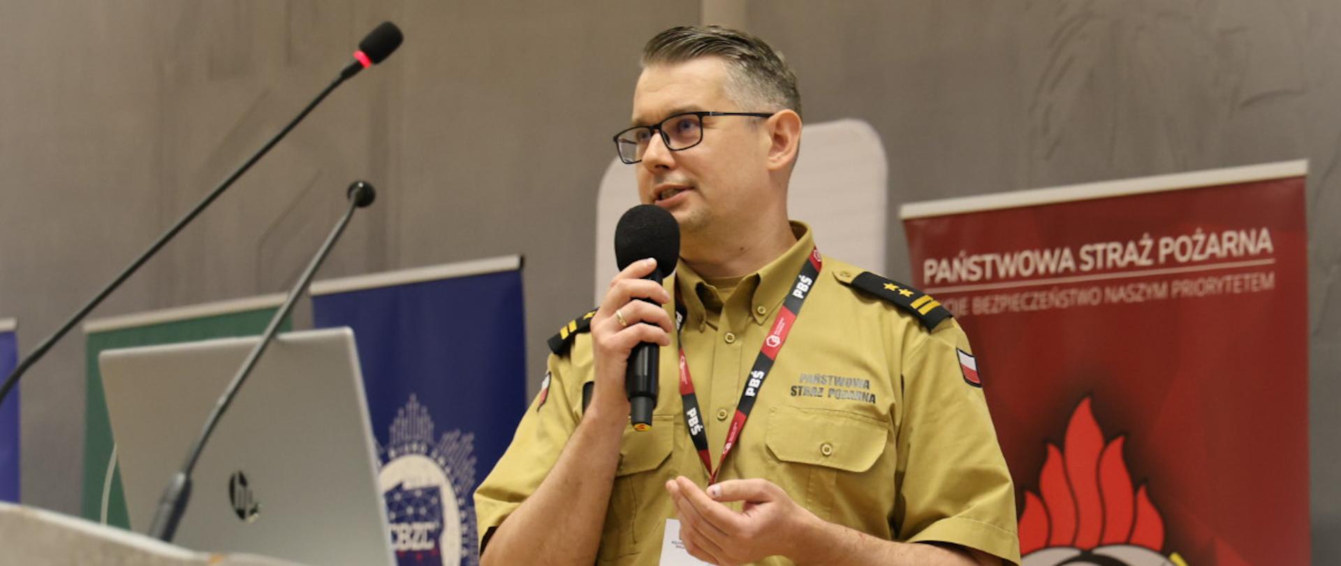 Mężczyzna w okularach i beżowym mundurze służbowym Państwowej Straży Pożarnej stoi za jasną mównicą, na której znajduje się otwarty laptop. W prawej ręce trzyma czarny mikrofon, a na szyi ma zawieszoną smycz z identyfikatorem z napisem „GOŚĆ”. Po prawej stronie w tle stoi czerwony baner z godłem Polski, logo straży oraz napisem „PAŃSTWOWA STRAŻ POŻARNA”. Po lewej stronie, za mównicą, widoczny jest niebieski baner z logo Centralnego Biura Zwalczania Cyberprzestępczości (CBZC).