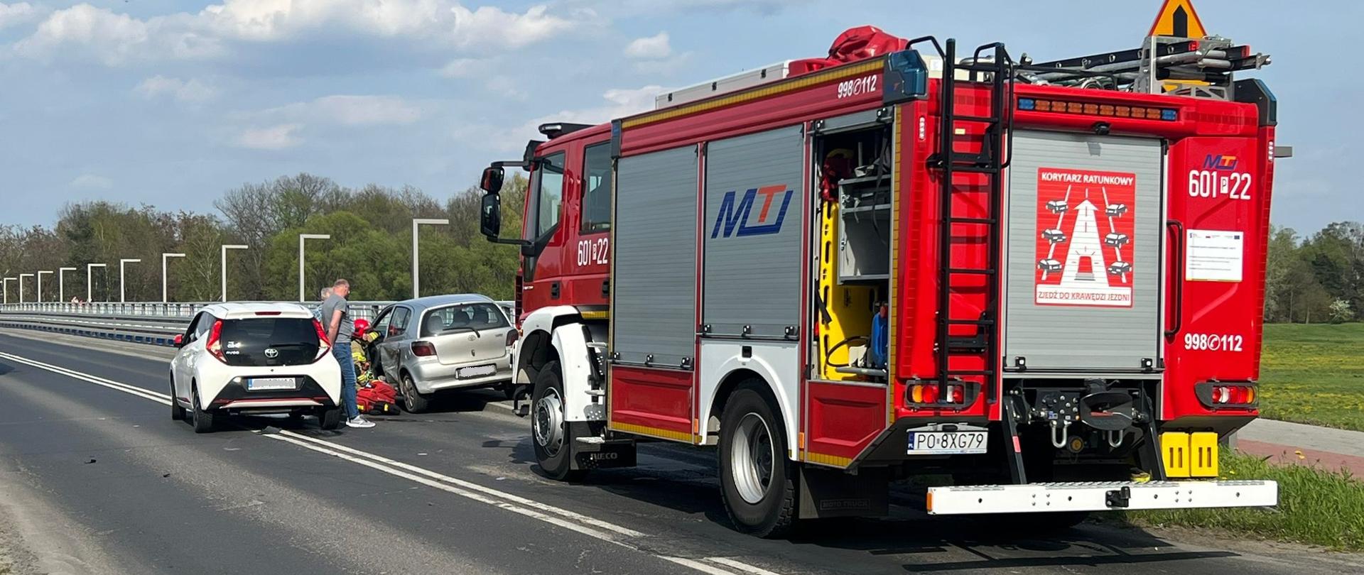 Na zdjęciu czerwony samochód strażacki, strażacy udzielający pomocy poszkodowanemu, za nimi wraki pojazdów