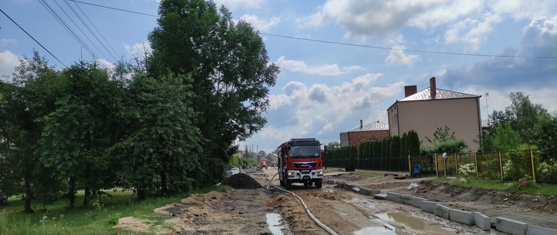 Teren budowy ulicy, rozkopane pobocza na których ustawione są materiały do budowy drogi, na drugim planie samochód pożarniczy z uruchomionymi światłami błyskowymi, z samochodu poprowadzona jest linia W-75 zasilająca kurtyny wodne