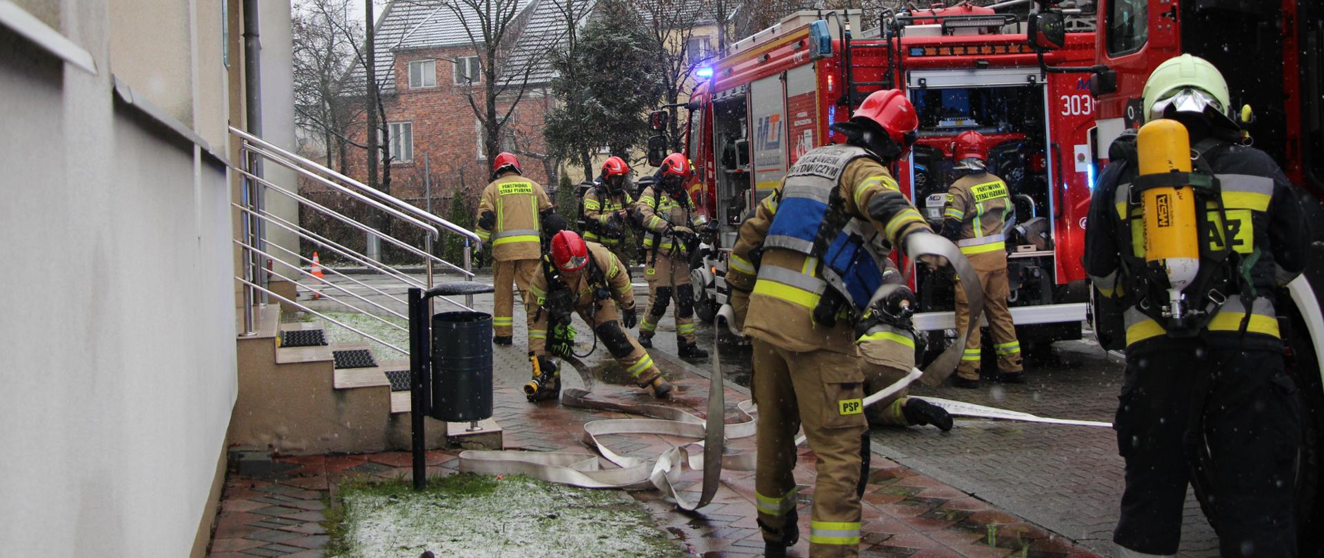 Zdjęcie przedstawia strażaków podczas rozwijania sprzętu przy wejściu do budynku jednego ze skrzydeł szpitalnych po prawej stronie na jezdni zaparkowały 2 wozy strażackie.