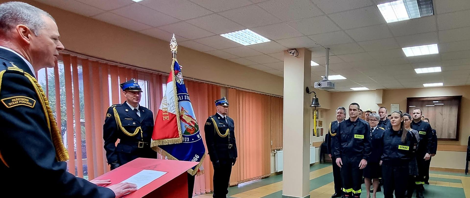 Na świetlicy szkoleniowej od prawej strony stoi komendant, w centrum kadru trzech umundurowanych strażaków w poczcie sztandarowym i po prawej stronie stoi grupka strażaków.