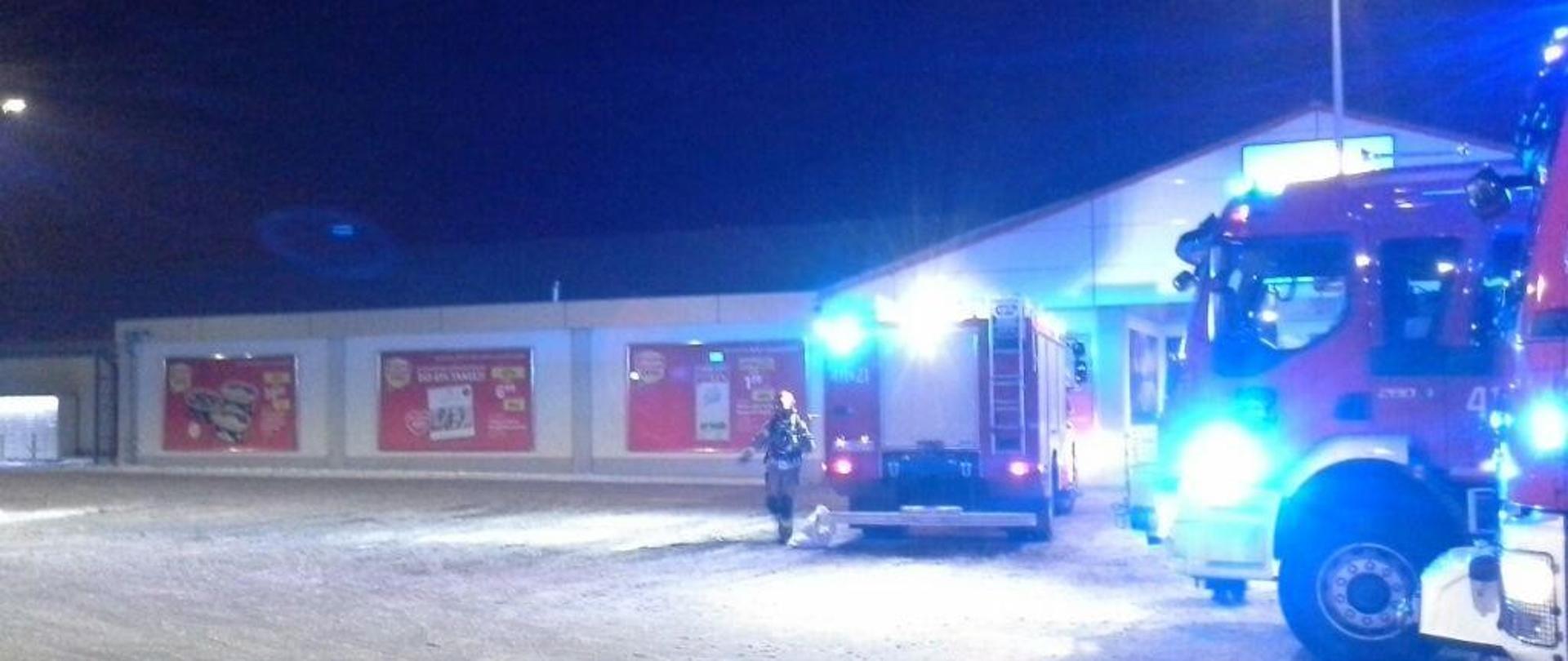 Parking obiektu handlowego, pora nocna. Widoczne samochody strażackie z włączonymi światłami błyskowymi w kolorze niebieskim. W tle jeden ze strażaków