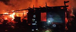 Na zdjęciu widoczny strażak w ubraniu specjalnym granatowym w hełmie białym przed płonącym drewnianym budynkiem mieszkalnym.