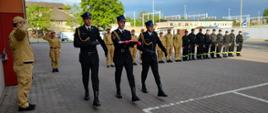 Z szeregu wymaszerował poczet flagowy, trzech strażaków w umundurowaniu reprezentacyjnym, flagowy na wyciągniętych rękach niesie biało-czerwoną flagę. Pozostali strażacy z szeregu są ubrani w piaskowe ubrania służbowe oraz czarne ubrania koszarowe. 