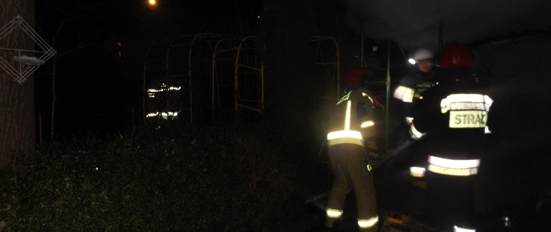 Zdjęcie przedstawia strażaków gaszących pożar altany. Jest ciemno. W tle widać żółtą pergolę.
