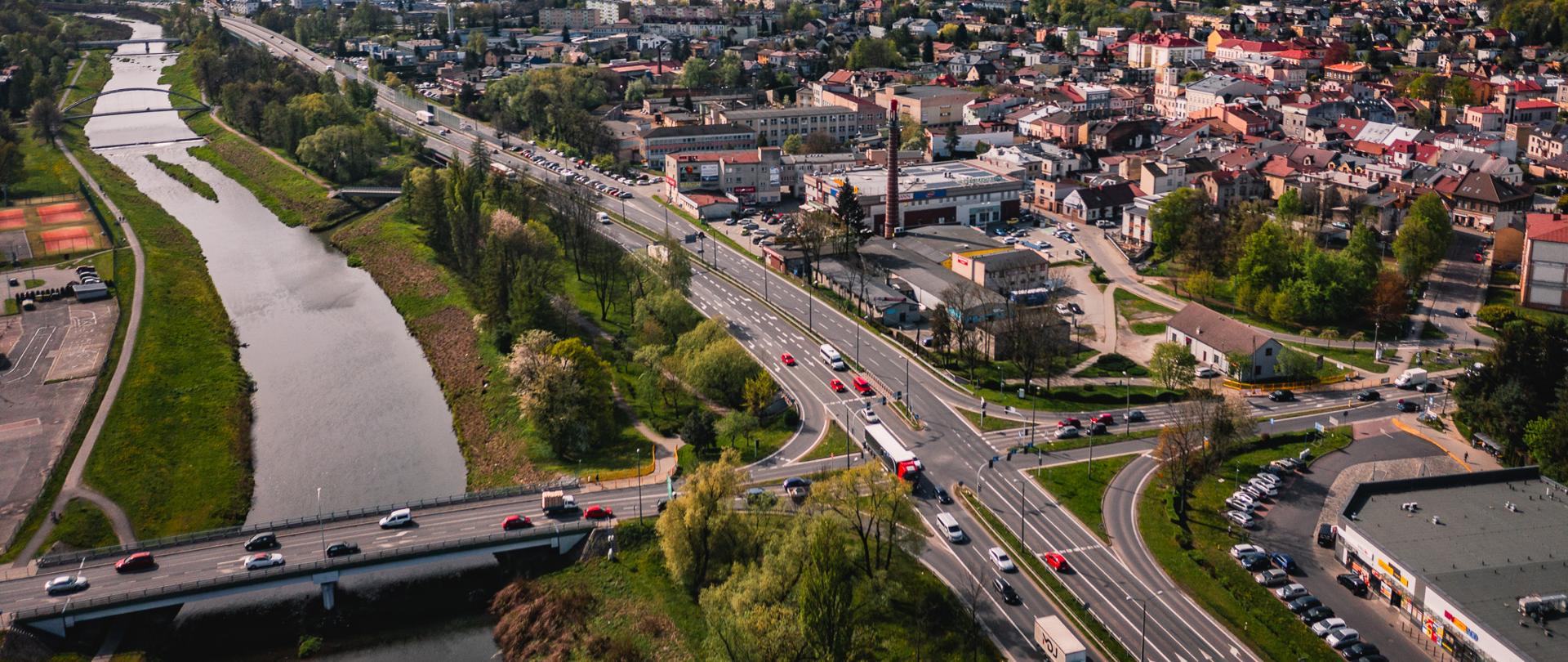 Panorama miasta Skoczów z lotu ptaka, widoczna droga krajowa DK81 biegnąca wzdłuż rzeki Wisły, most nad rzeką, zabudowa miejska oraz otaczające tereny zielone, w tle pasmo górskie Beskidu Śląskiego.