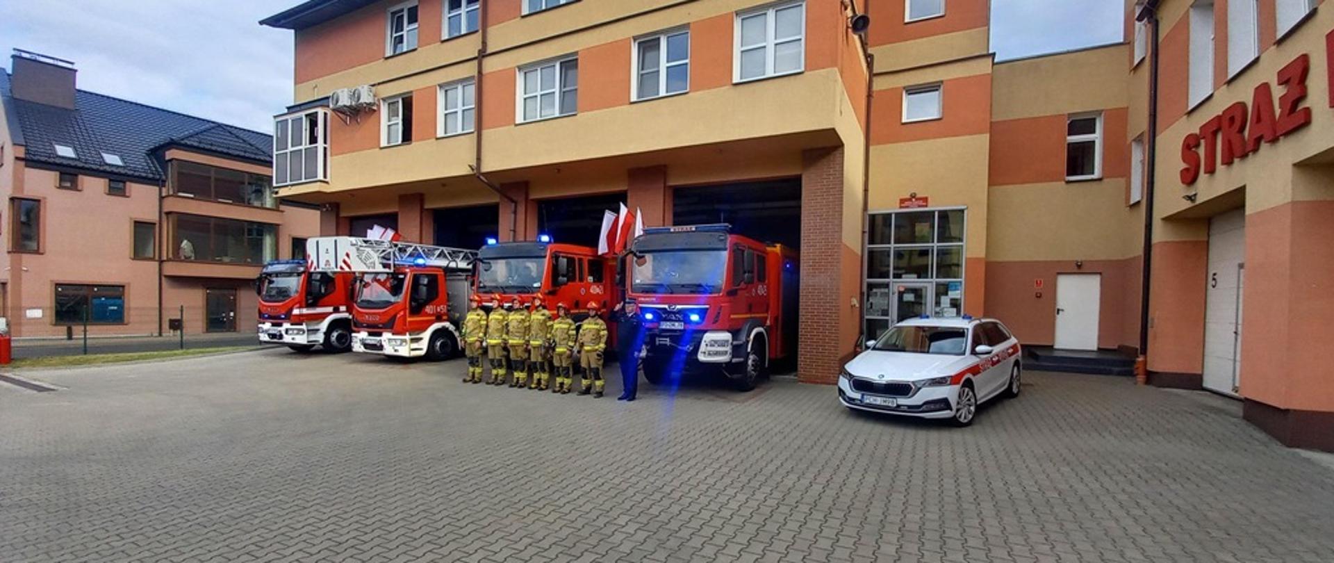Zdjęcie przedstawia strażaków stojących w szeregu na tle budynku komendy i samochodów pożarniczych. 