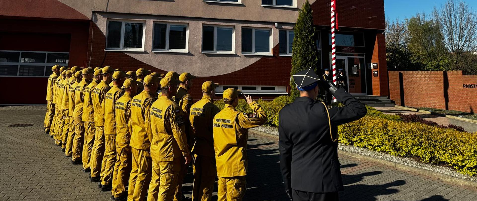 Na zewnątrz, przed budynkiem Państwowej Straży Pożarnej, stoi w szeregu grupa strażaków w piaskowym umundurowaniu służbowym. Są oni zwróceni w stronę masztu. Na pierwszym planie, po prawej stronie, widoczny jest od tyłu funkcjonariusz w ciemnym, wyjściowym mundurze, który salutuje w stronę biało-czerwonego masztu. W tle znajduje się dwupiętrowy, ceglasto-beżowy budynek oraz bezchmurne, jasnoniebieskie niebo.