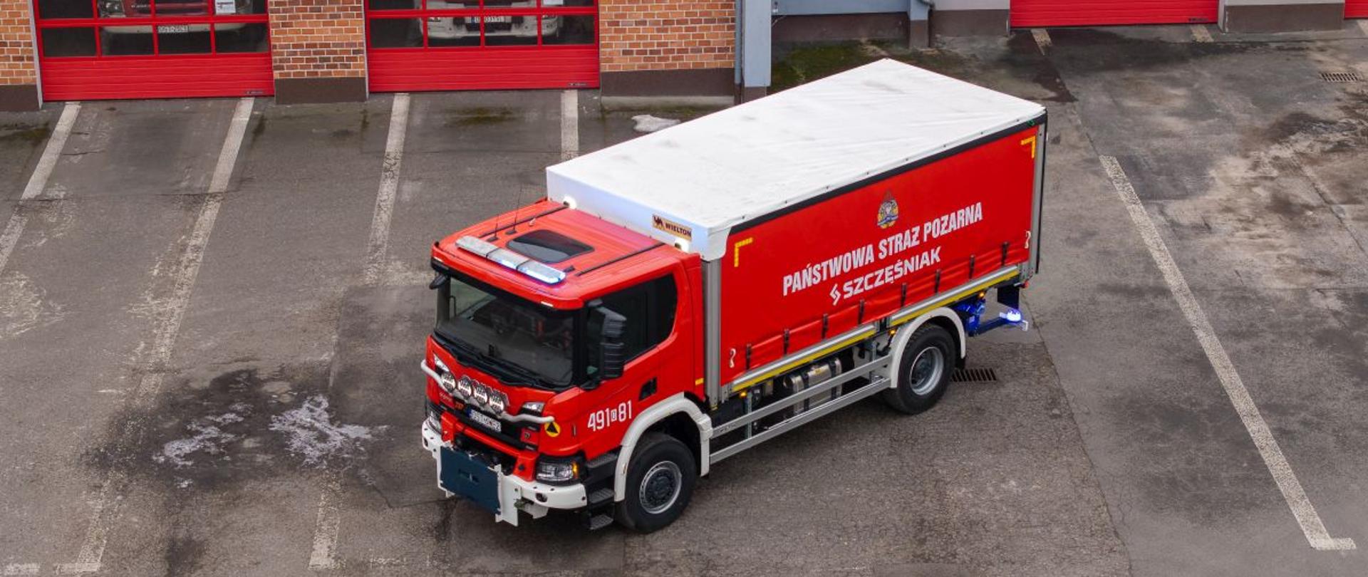 Czerwony samochód kwatermistrzowski Państwowej Straży Pożarnej stoi przed budynkiem strażnicy z kilkoma bramami garażowymi. Na burcie widoczne są oznaczenia PSP i numer 491[O]81. Pojazd ma białą górną część kabiny i wyposażenie charakterystyczne dla pojazdów logistycznych.