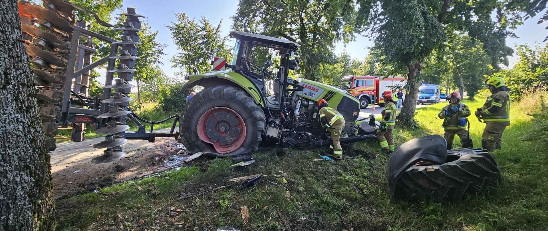 Na zdjęciu widoczny jest poważny wypadek z udziałem ciągnika rolniczego. Zielony ciągnik marki CLAAS z podczepioną maszyną rolniczą znajduje się częściowo poza jezdnią, przy drzewie. Jego przednia część jest mocno uszkodzona, a jedno z dużych kół leży oderwane na trawie obok pojazdu. Wokół ciągnika znajdują się porozrzucane fragmenty elementów pojazdu Na miejscu pracuje kilku strażaków w ubraniach ochronnych i hełmach, którzy prowadzą działania przy uszkodzonym ciągniku. W tle, na wąskiej drodze otoczonej drzewami i zielenią, stoją samochody straży pożarnej oraz inne pojazdy służb ratunkowych.