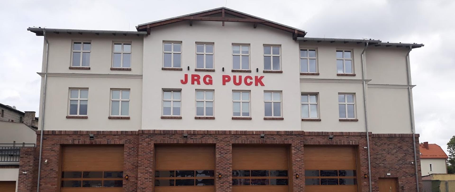 Zdjęcie przedstawia trzy kondygnacyjny budynek, na samym dole widać cztery brązowe bramy garażowe, powyżej dwa rzędy okien pomiędzy którymi znajduje się napis JRG Puck