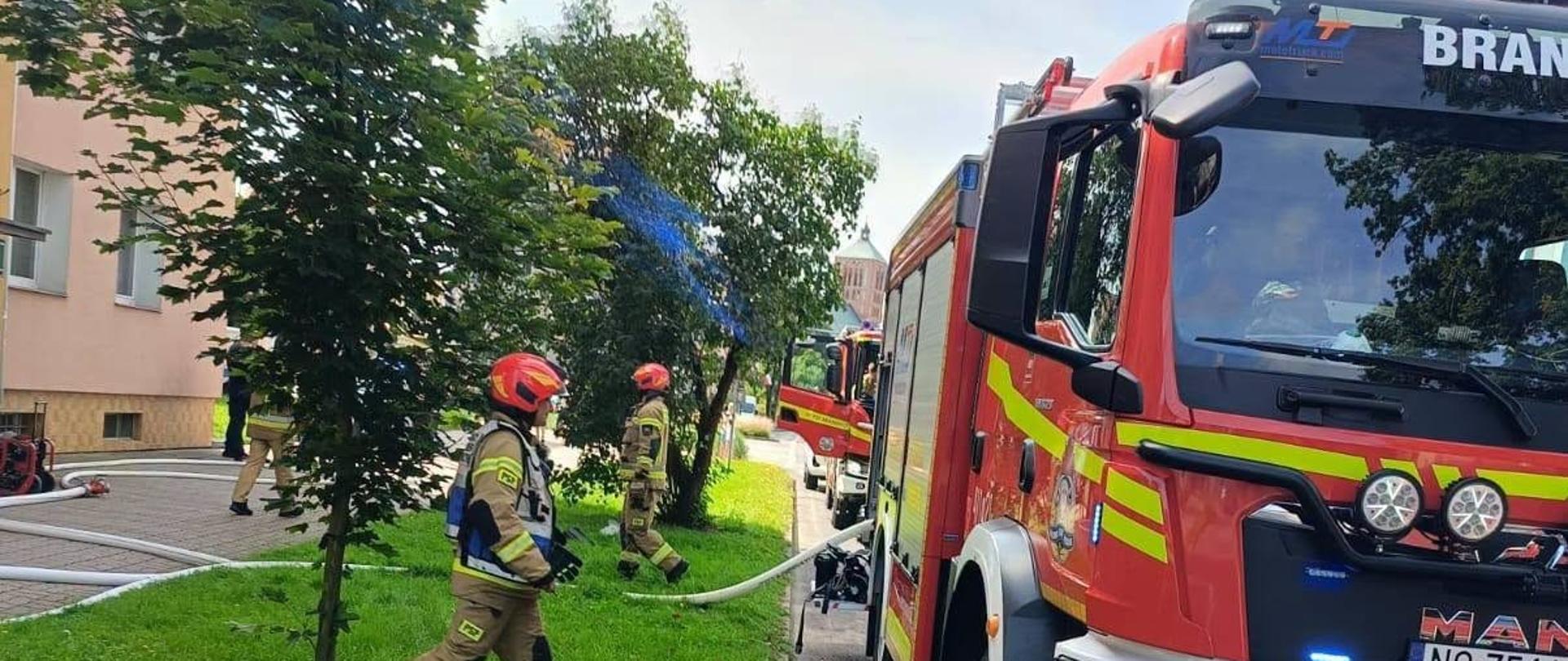 Na zdjęciu widoczna jest akcja straży pożarnej przy budynku mieszkalnym. Strażacy prowadzą działania z wykorzystaniem rozwiniętych linii wężowych, a na miejscu znajdują się co najmniej dwa samochody ratowniczo-gaśnicze. Jeden z pojazdów ma włączone niebieskie światła alarmowe.