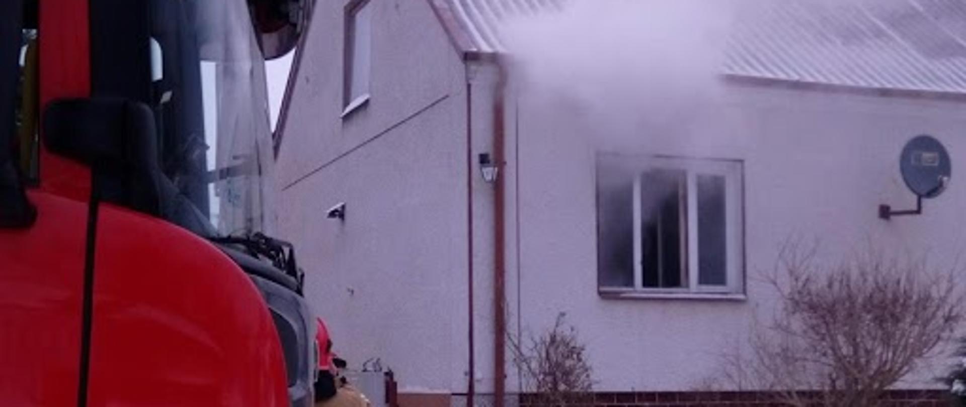 Zdjęcie przedstawia przód kabiny samochodu gaśniczego stojącego na zaśnieżonej drodze przed wjazdem na posesję, na drugim planie biały budynek mieszkalny z czerwonym dachem z widocznymi kłębami białego dymu wydobywającego się z otwartych okien jednego z pomieszczeń budynku. 
