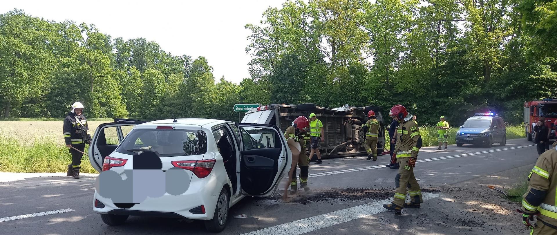 Na pierwszym planie widać biały samochód osobowy, który ma otwarte drzwi i stoi na drodze. W tle widać biały samochód dostawczy, który leży na lewym boku na jezdni. Obok widać strażaków, policjanta oraz osoby postronne. W oddali widać czerwony samochód straży pożarnej, jeden radiowóz policji, zielony znak z napisem Stara Sobótka 1 oraz drzewa. Jest widno.