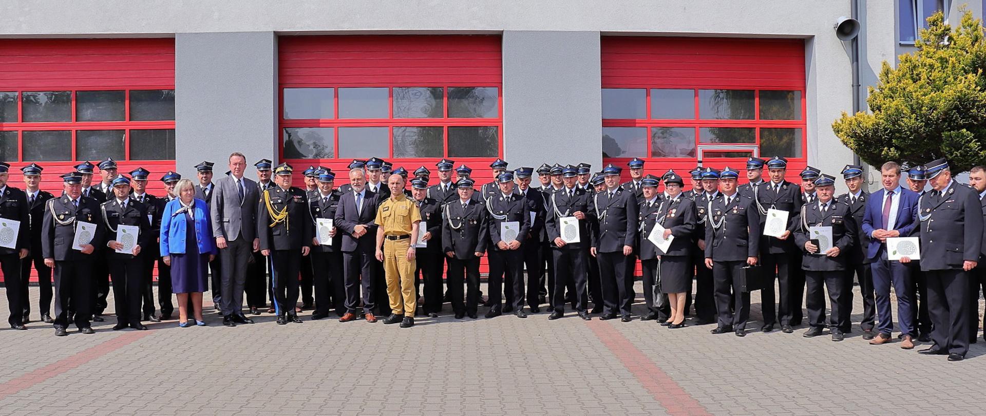 druhowie osp strażacy i zaproszeni goście pozują do zdjęcia na tle bram garażowych jednostki ratowniczo-gaśniczej