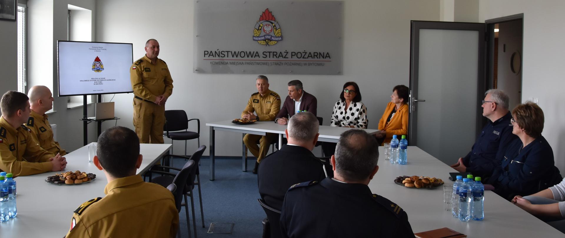 Na zdjęciu widoczna sala. Przy białych Stołach siedzą uczestnicy narady. Na ścianie w tle widoczne logo Państwowej Straży Pożarnej. Jedna z osób stoi i przemawia do reszty.
