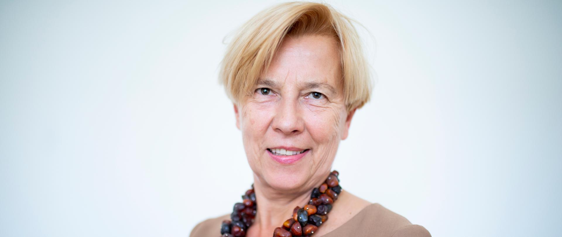 Na białym tle zdjęcie minister Małgorzaty Zielińskiej. Minister ubrana jest w beżową bluzkę i korale