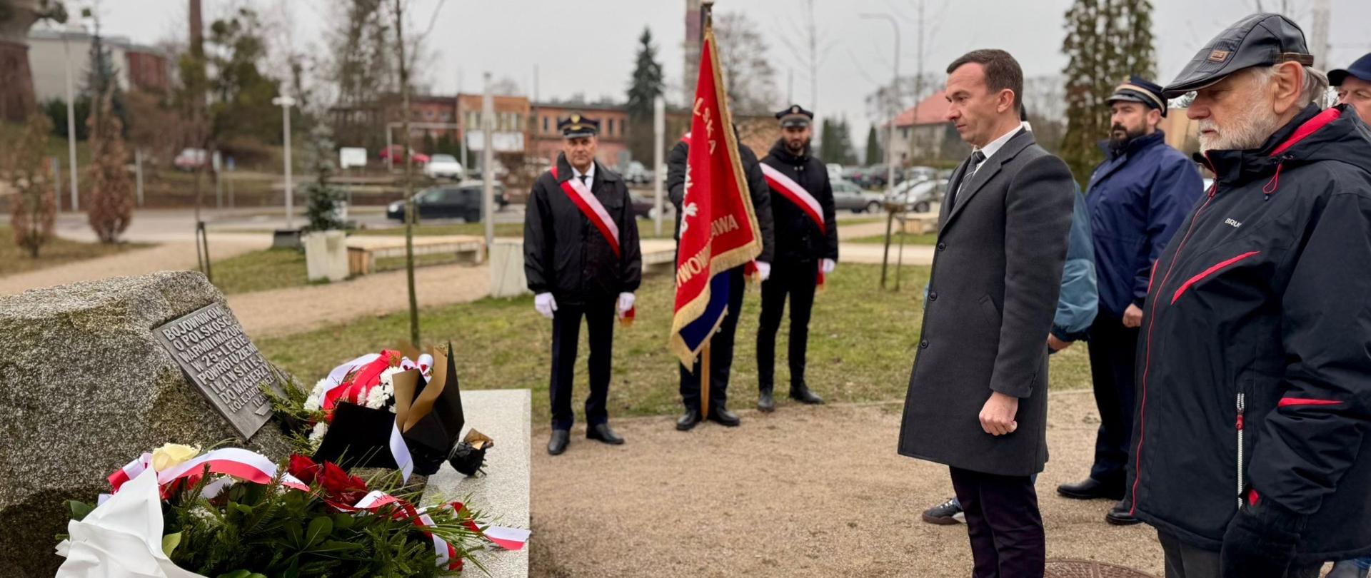 Mężczyzna w czarnym płaszczu stoi na baczność pod pomnikiem na Placu Iławskich Kolejarzy w Iławie. 