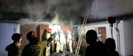 Na zdjęciu widzimy strażaków biorących udział w gaszeniu pomieszczenia gospodarczego. Strażacy ubrani są w odzież ochronną koloru piaskowego, na głowie czerwone hełmy, na plecach butle aparatu ochrony dróg oddechowych. Z wejścia do pomieszczenia buchają płomienie oraz wydobywa się czarny dym. Akcja gaśnicza oświetlana jest reflektorem. 