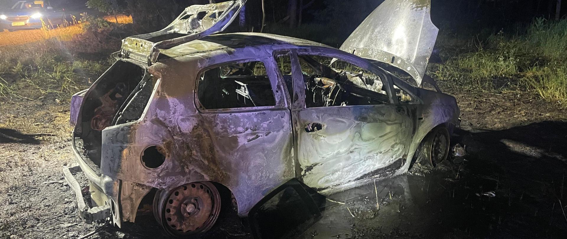 Na zdjęciu widoczny spalony samochód osobowy, całkowicie zwęglony i zniszczony, stoi na poboczu drogi. Jego maska i klapa bagażnika są otwarte, a wnętrze jest doszczętnie spalone. W tle widać fragment drogi, las i światła innego pojazdu.