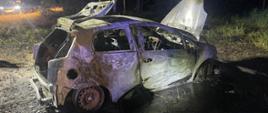 Na zdjęciu widoczny spalony samochód osobowy, całkowicie zwęglony i zniszczony, stoi na poboczu drogi. Jego maska i klapa bagażnika są otwarte, a wnętrze jest doszczętnie spalone. W tle widać fragment drogi, las i światła innego pojazdu.
