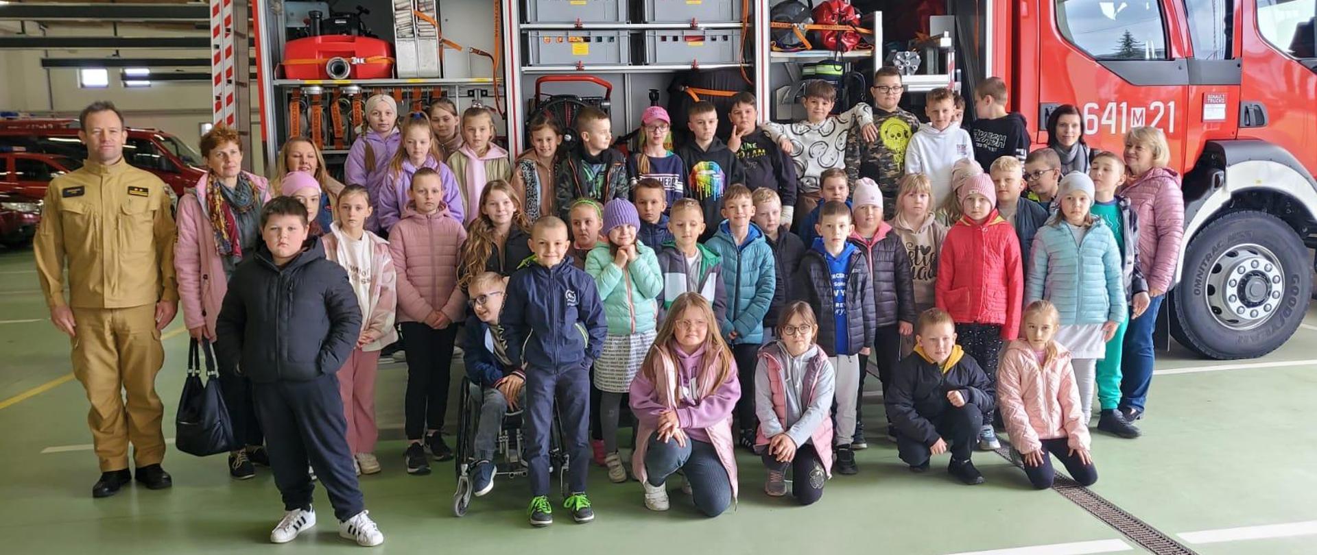 Kolejne odwiedziny strażnicy i sali edukacyjnej typu „Ognik” w KP PSP w Sokołowie Podlaskim 19.04.2024 r. - zdjęcie grupowe w garażu w tle samochód strażacki