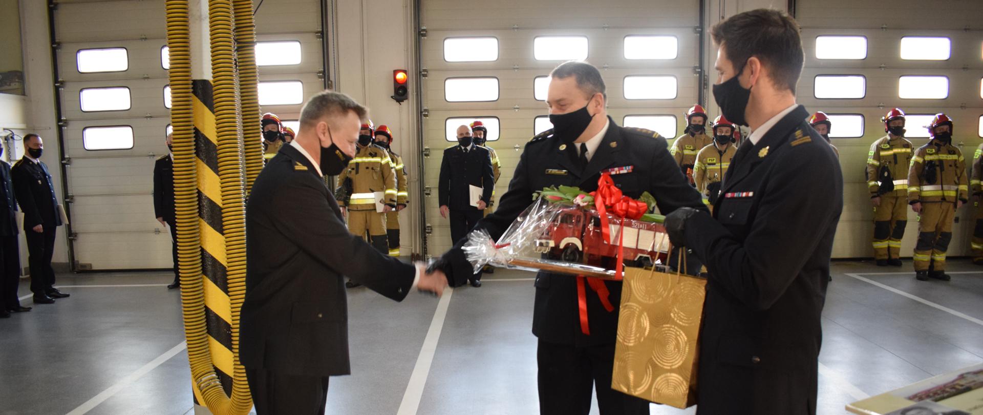Na pierwszym planie znajdują się 3 mężczyźni, zwróceni bokiem/profilem do fotografa. Jeden znajduje się po lewej stronie, a dwóch na przeciw niego po prawej stronie, wręczają mu prezent. Jeden mężczyzna z prawej ściska dłoń temu stojącemu po lewej stronie. Wszyscy 3 mężczyźni ubrani są w mundur strażacki galowy. Na drugim planie widać ustawionych w dwuszeregu strażaków w pełnym umundurowaniu. Wszyscy znajdują się w garażu strażnicy komendy.