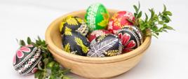 Na białym tle drewniana miseczka a w niej pisanki wielkanocne. Po lewej stronie zielona gałązka bukszpanu i pisanka.