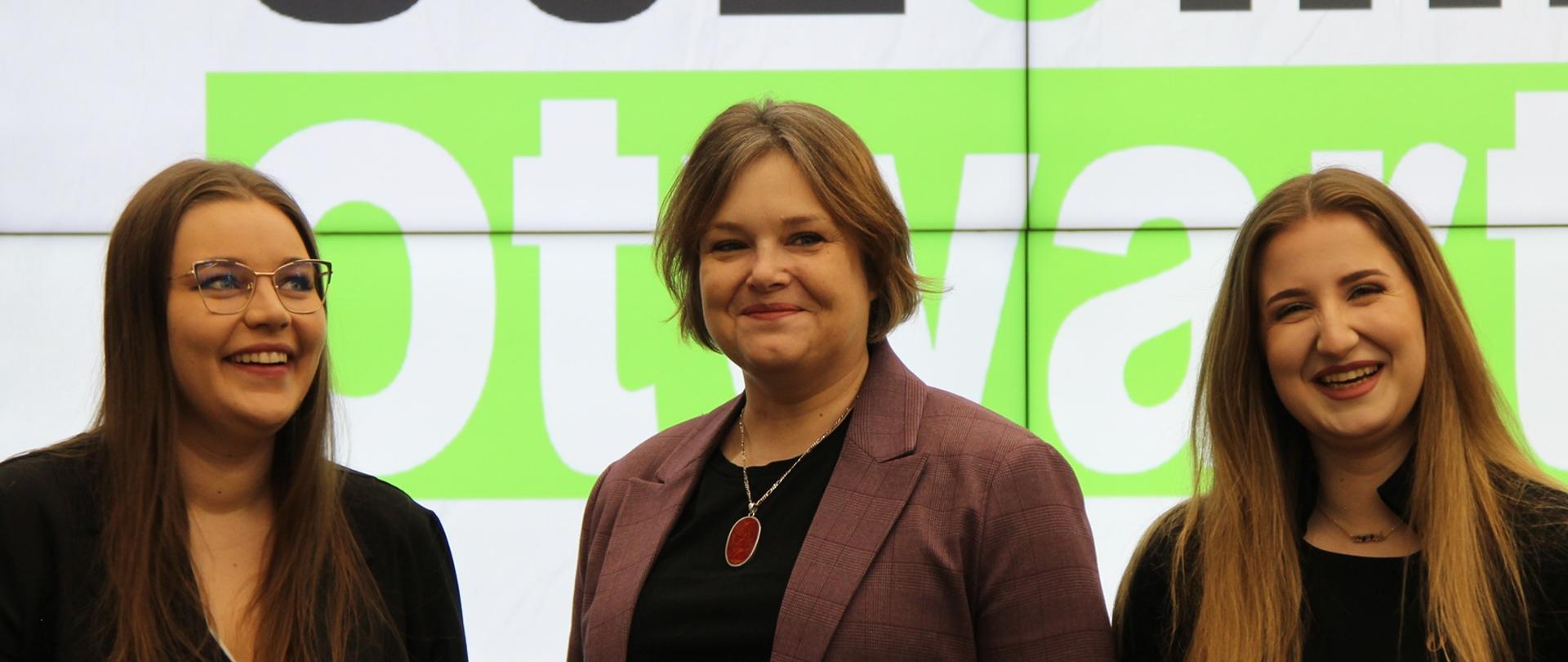 Wiceminister Zioło-Pużuk stoi między dwoma młodymi ubranymi na czarno dziewczynami, za nimi na wielkim ekranie biały napis Uczelnia otwarta na zielonym tle.