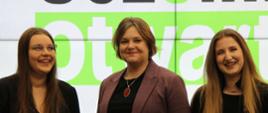 Wiceminister Zioło-Pużuk stoi między dwoma młodymi ubranymi na czarno dziewczynami, za nimi na wielkim ekranie biały napis Uczelnia otwarta na zielonym tle.