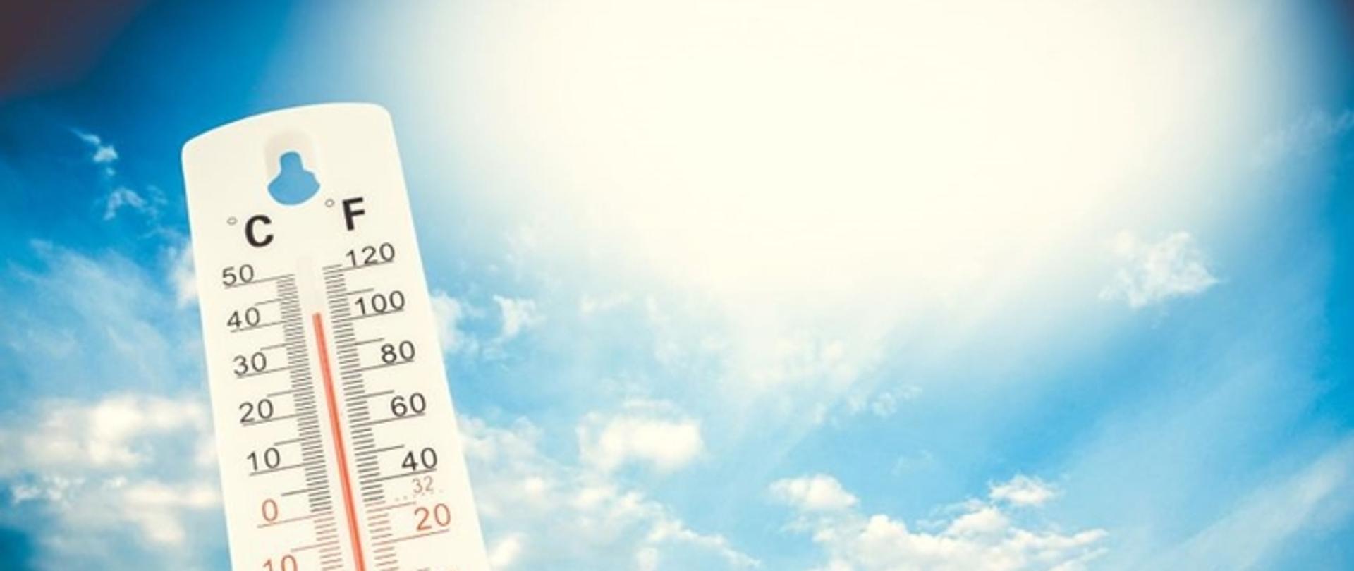 Obraz przedstawiający termometr który wskazuje wysoką temperaturę na tle słonecznego nieba.