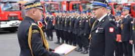 Zdjęcie zrobione na zewnątrz podczas uroczystości. Oficer Państwowej Straży Pożarnej w stopniu generała wręcza funkcjonariuszowi Państwowej Straży Pożarnej oprawiony w ramki dyplom. Za nimi stoją ustawieni strażacy Państwowej Straży Pożarnej wraz z pocztem sztandarowym. W tle stoją ustawione samochody ratowniczo-gaśnicze. 